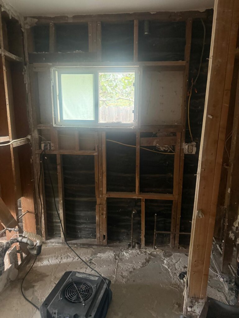 Bathroom wall opened to the studs below a window, with an air mover drying the area